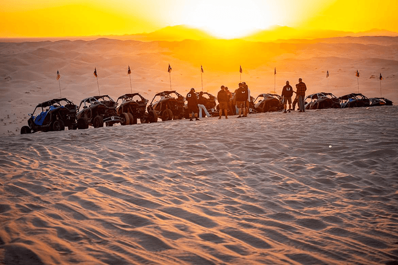 Riders and side-by-sides gathered along a dune at sunset